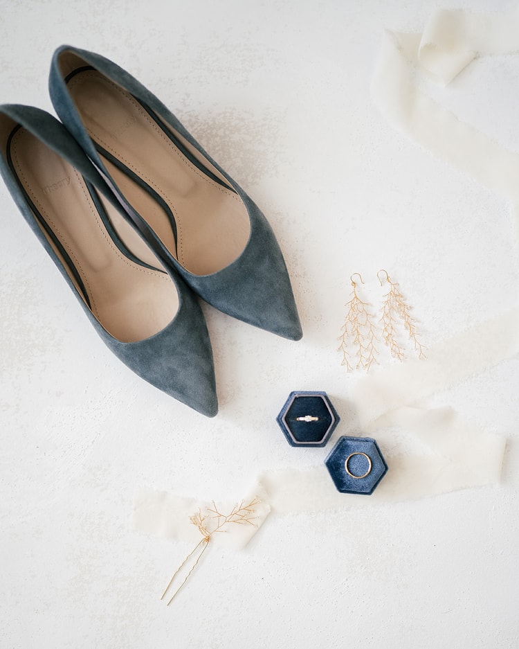 Flat-lay of bridal details: dusty blue suede heels, gold branch earrings, a hair pin, and wedding rings in navy velvet hexagonal boxes on a white surface.