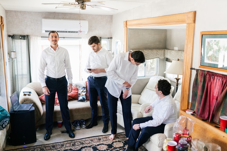 Groomsmen helping each other dress in white button-down shirts and navy trousers inside a casual getting-ready room.