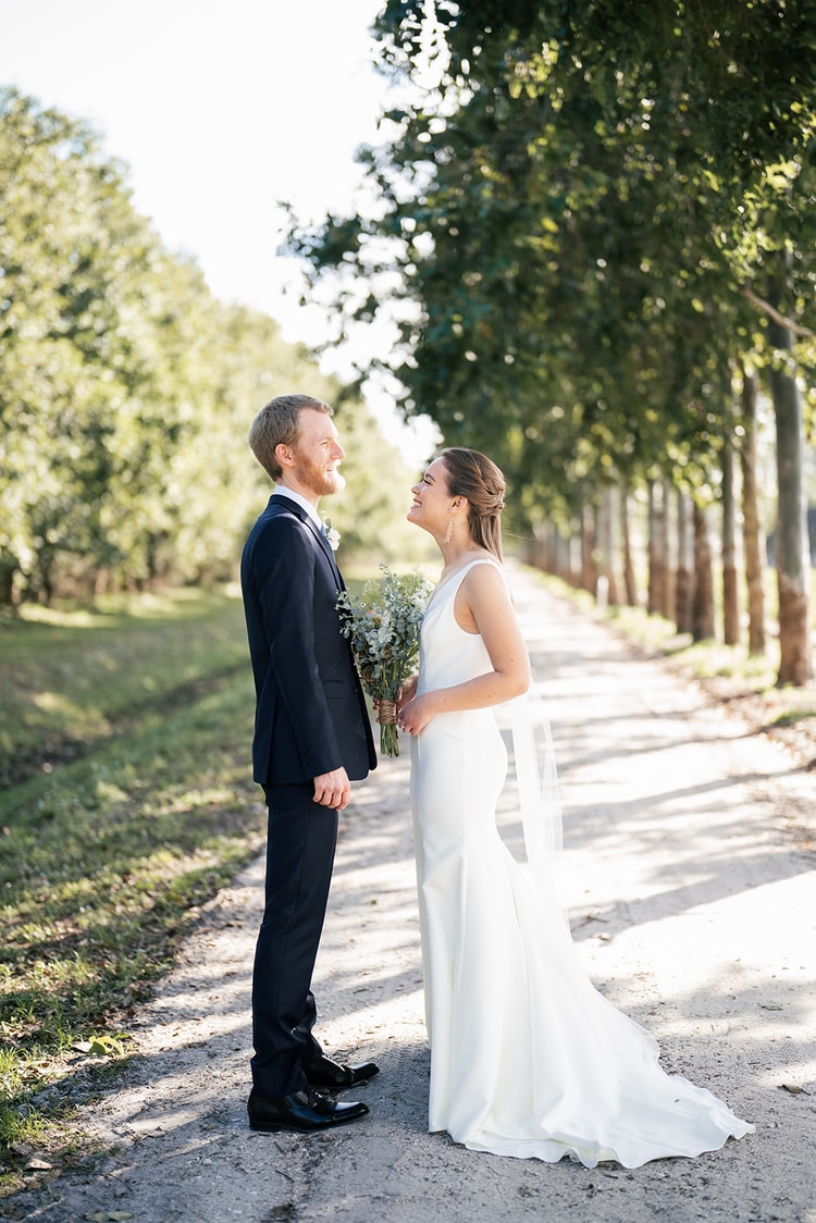 Bride and groom standing face to face along a tree-lined lane, smiling up at each other in golden afternoon light.