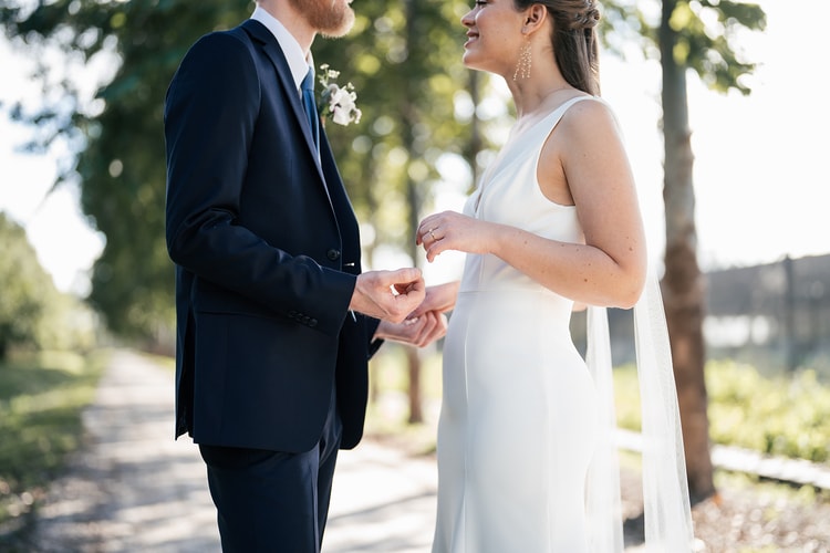 Couple holding hands and sharing a look during outdoor portraits, the bride's veil trailing gently beside them in a sunlit grove.