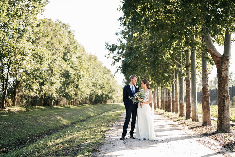 Bride and groom standing together on a tree-lined path during portraits, facing each other and smiling in warm afternoon light.