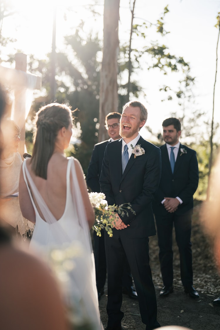 Groom breaking into a wide laugh as the bride approaches the altar during the outdoor ceremony, groomsmen looking on.