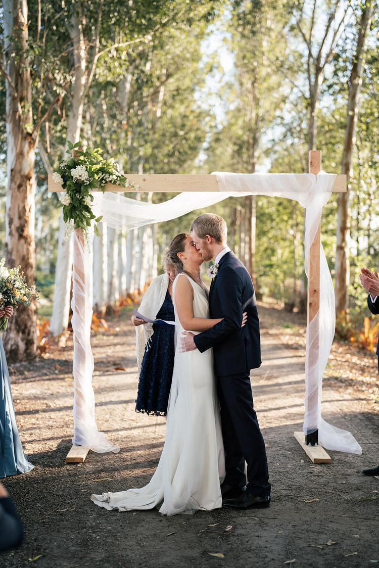 First kiss at the altar under a draped wooden arch, with guests applauding on either side during the outdoor ceremony.