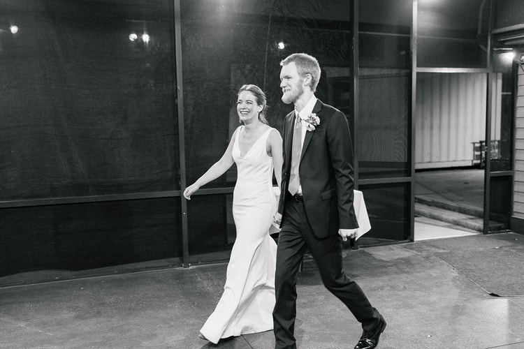 Black-and-white photo of the newlywed couple walking hand in hand and laughing as they enter their reception venue.