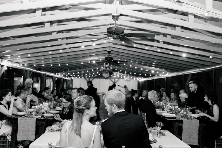 Black-and-white reception scene: couple seated at the sweetheart table in the foreground as a guest gives a speech to the room.