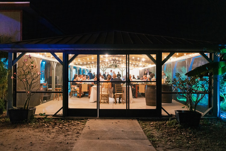 Wide nighttime view from outside the glowing glass pavilion, showing guests seated at long tables under warm string lights.