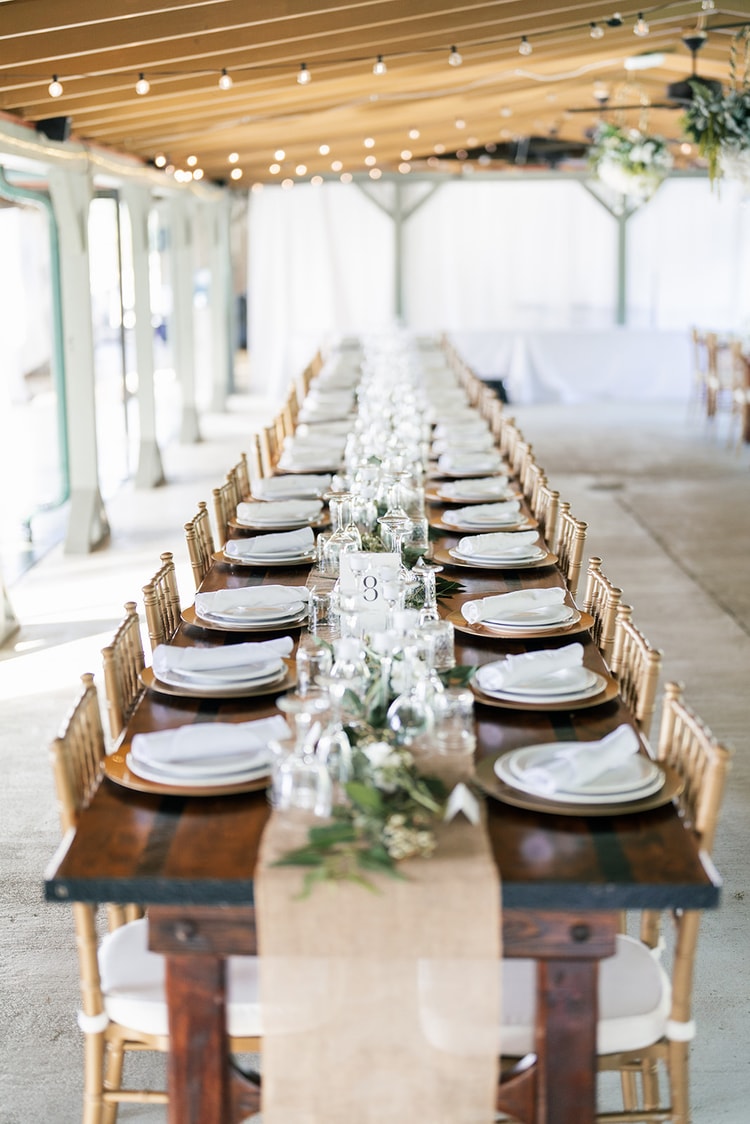 Long reception table set with white plates, gold chargers, greenery runners, and glass votives under café lights in a covered pavilion.