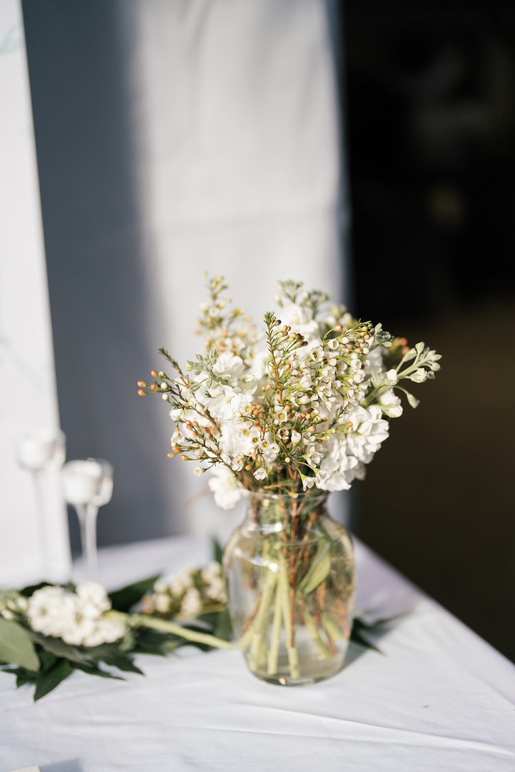 Glass vase of white stock and waxflower blooms on a draped sweetheart table, part of the reception floral decor.