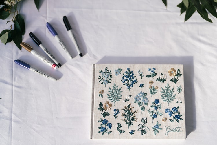 Floral guest book with a botanical embroidered cover on a white-draped table, surrounded by markers, ready for guests to sign.