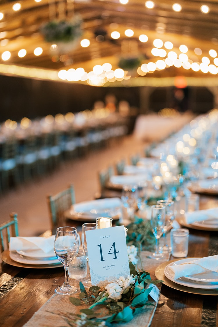 Table number 14 card at a warmly lit reception table, with greenery runners and gold charger plates stretching back under a canopy of Edison bulbs.