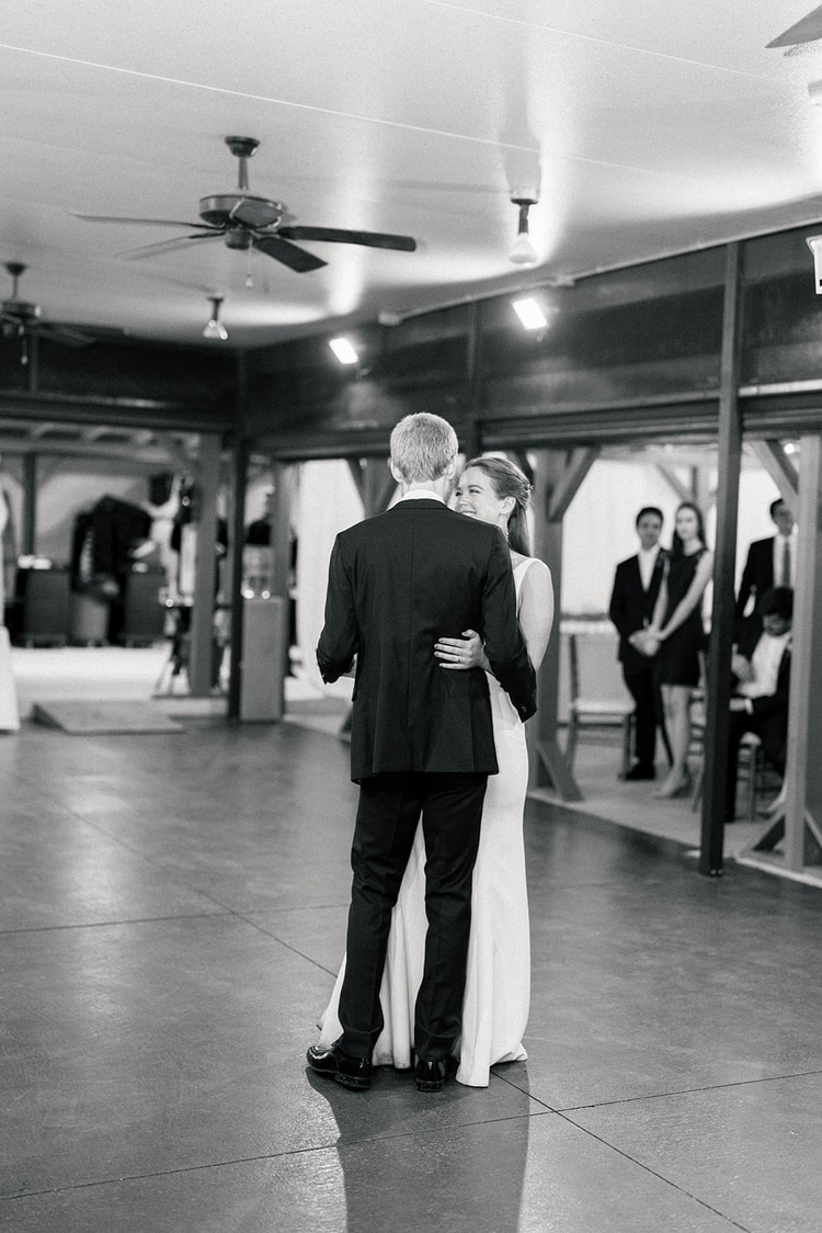 Black-and-white first dance, the couple swaying with their backs to camera, the bride smiling over his shoulder at onlookers.