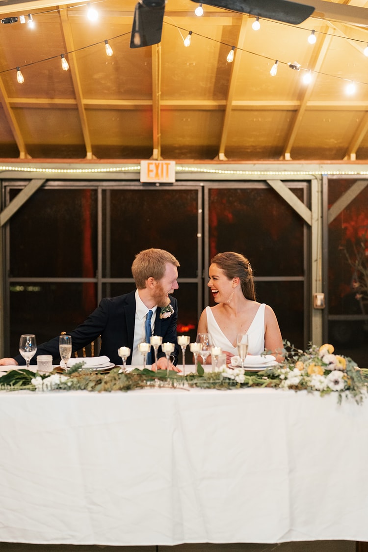 Couple at the sweetheart table laughing with each other, surrounded by candlelight and an anemone-and-greenery floral runner.