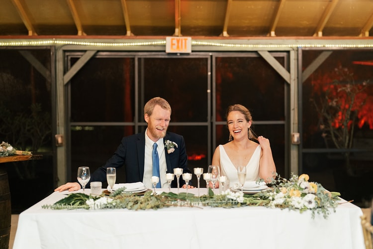Couple laughing together at the sweetheart table during speeches, floral greenery and candles spread across the table in front of them.