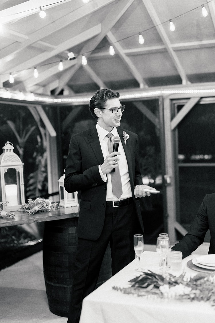 Black-and-white portrait of a groomsman delivering a speech at the reception, microphone in hand, smiling warmly under string lights.