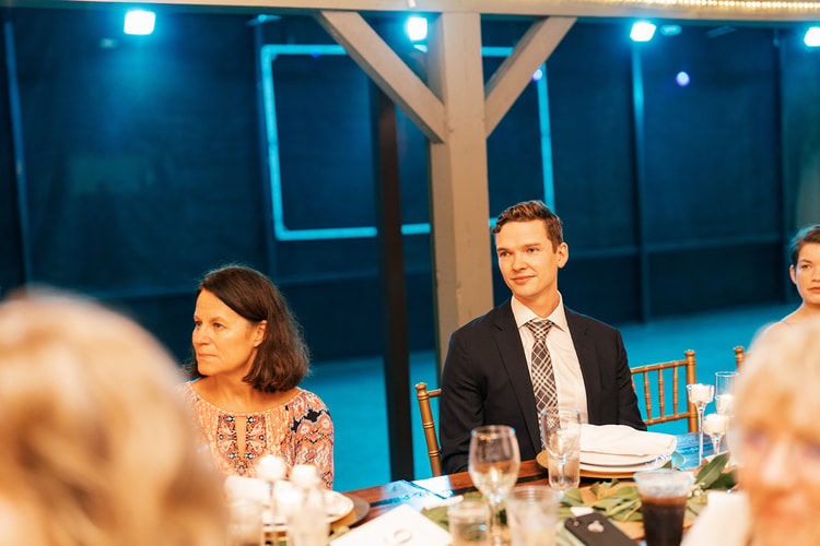 Wedding guests seated at a reception dinner table listening attentively during speeches, lit by blue ambient light.
