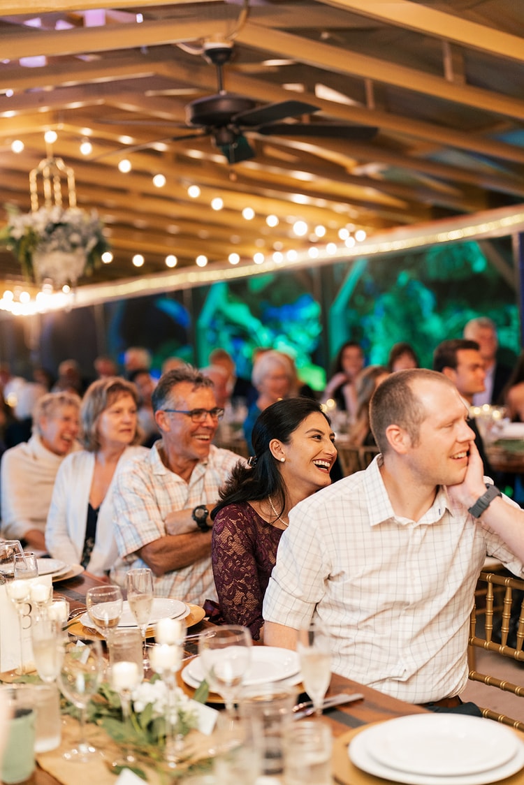 Reception guests laughing at their table under warm string lights, couples and families sharing a joyful moment during dinner.
