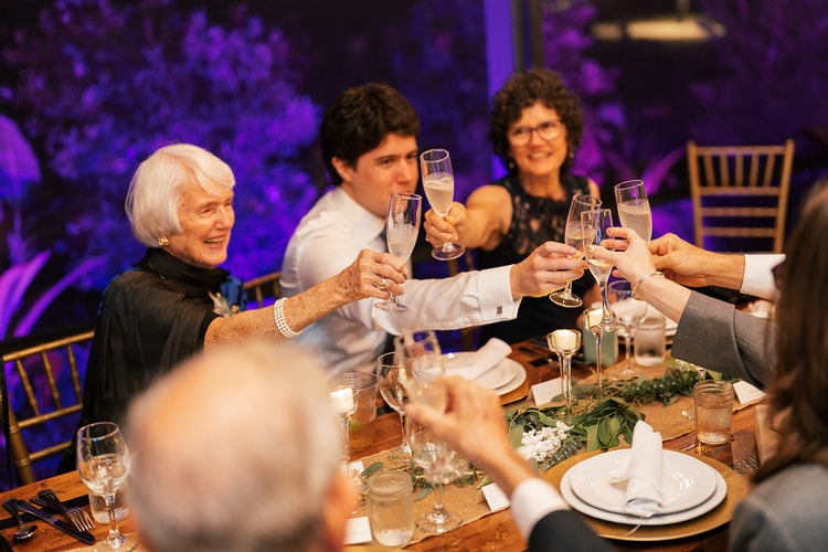 Reception guests clinking champagne flutes in a toast across a dinner table, three generations sharing a joyful moment.