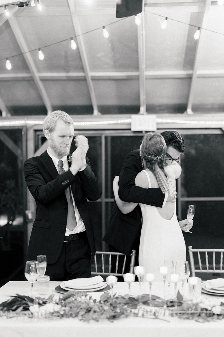 Black-and-white reception moment: groom wiping away tears while a guest embraces the bride, all three moved by a heartfelt speech.