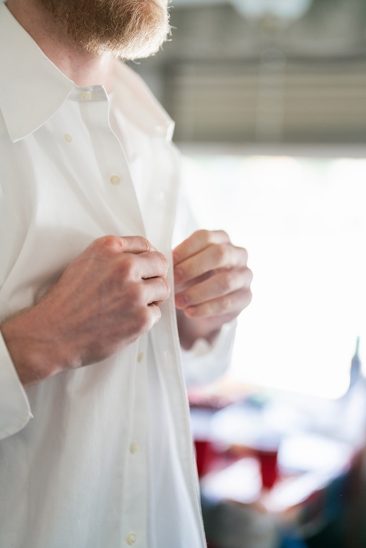Groom buttoning his white dress shirt, a close crop on his hands and chest, during morning preparations.