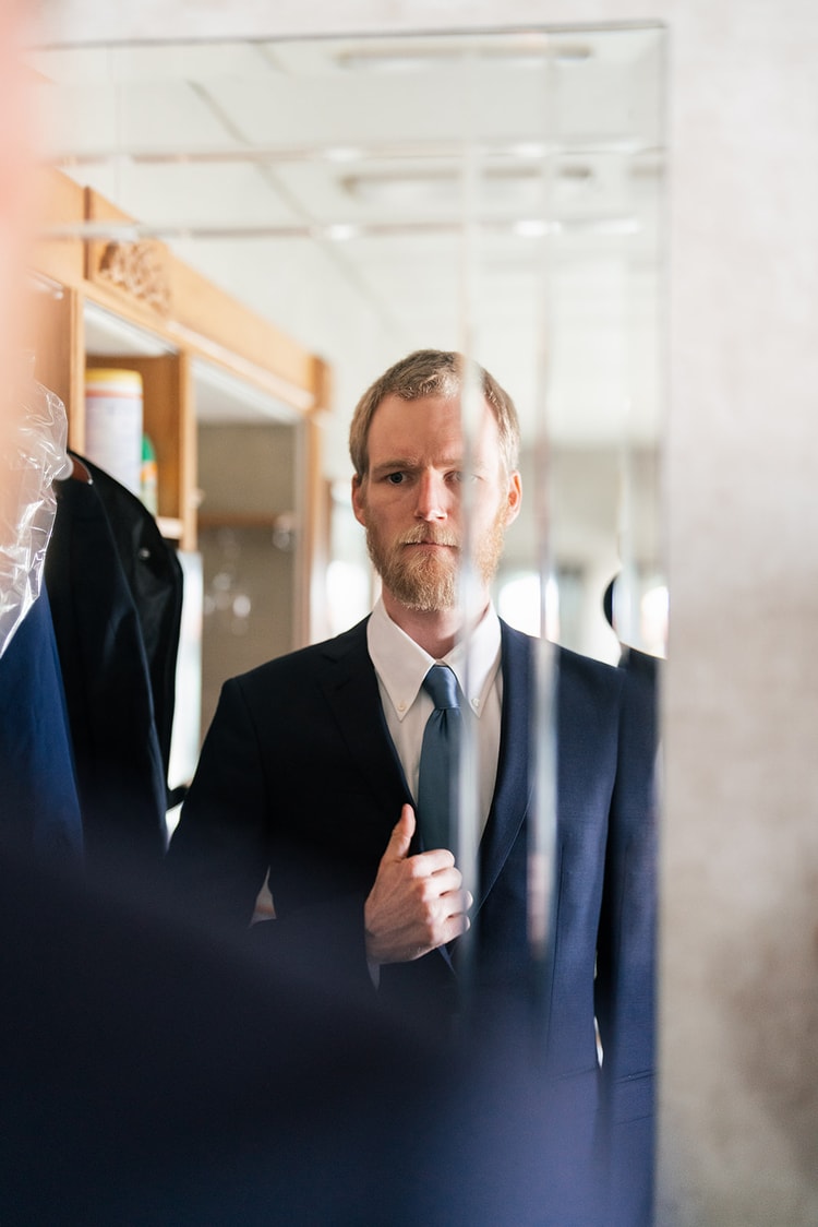 Groom in a navy suit and blue tie checking his reflection in a mirror while straightening his jacket before the ceremony.