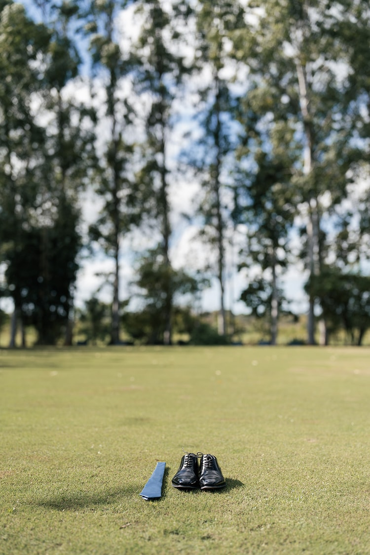 Groom's black dress shoes and blue tie arranged on a grass lawn with tall trees in the background.