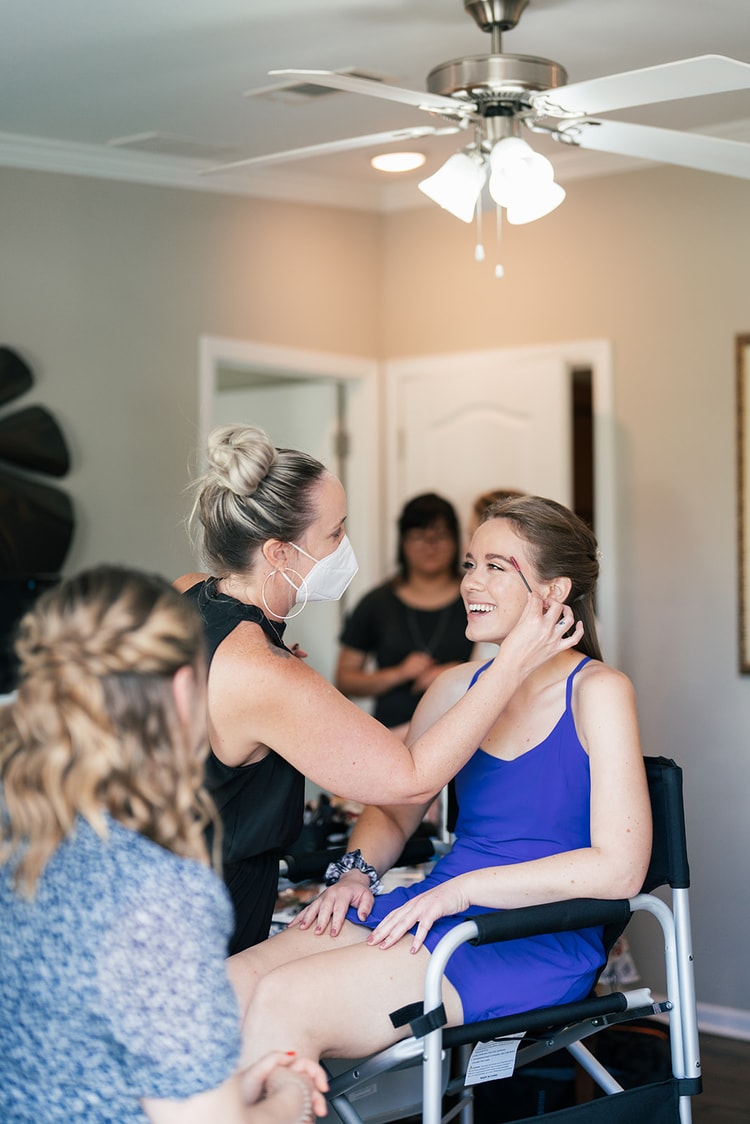 Makeup artist applying finishing touches to the smiling bride's face while a bridesmaid is attended to in the background during getting-ready preparations.