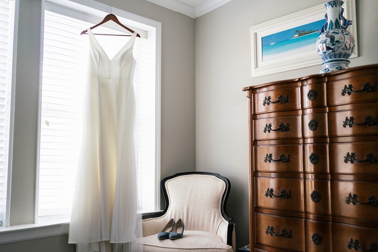 Wedding dress hanging in a sunlit window beside blue suede heels resting on a cream armchair, with an antique dresser and framed art nearby.