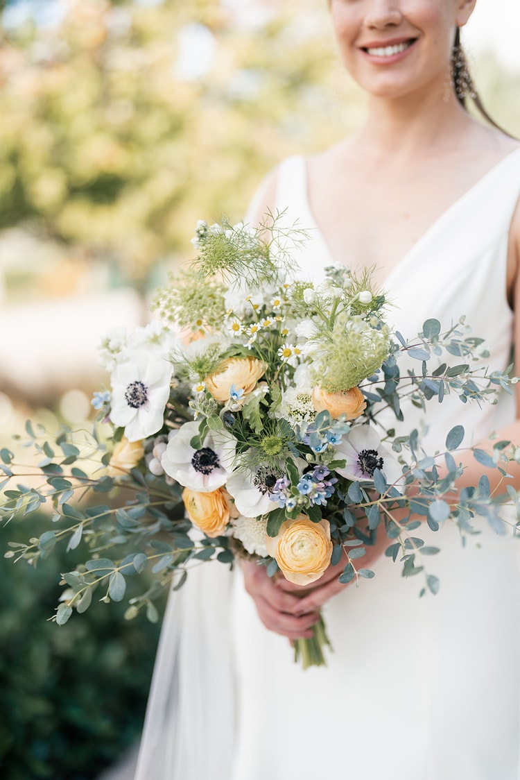 Close-up of the bride's bouquet featuring white anemones, peach ranunculus, blue wildflowers, chamomile, and cascading eucalyptus.