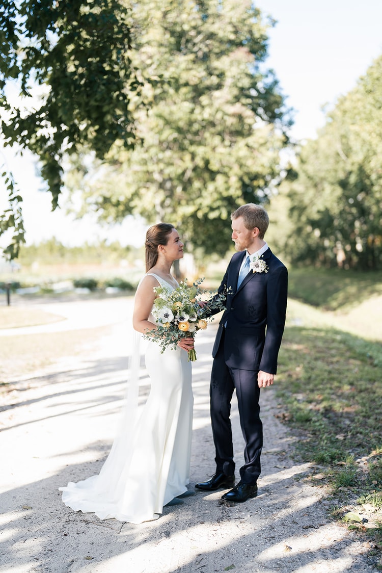 Bride and groom face each other on a sandy path lined with trees, sharing a quiet moment during couple portraits.