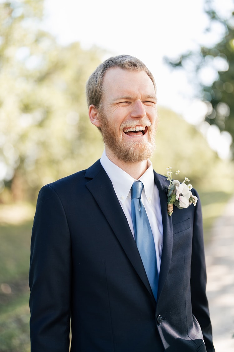 Groom in a navy suit and boutonniere laughing openly with his eyes squeezed shut, shot against a soft outdoor background.