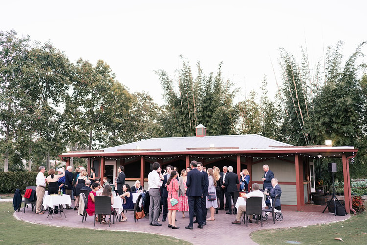 Wide exterior shot of a glass-walled reception pavilion glowing with string lights at night, guests gathered inside at long banquet tables.