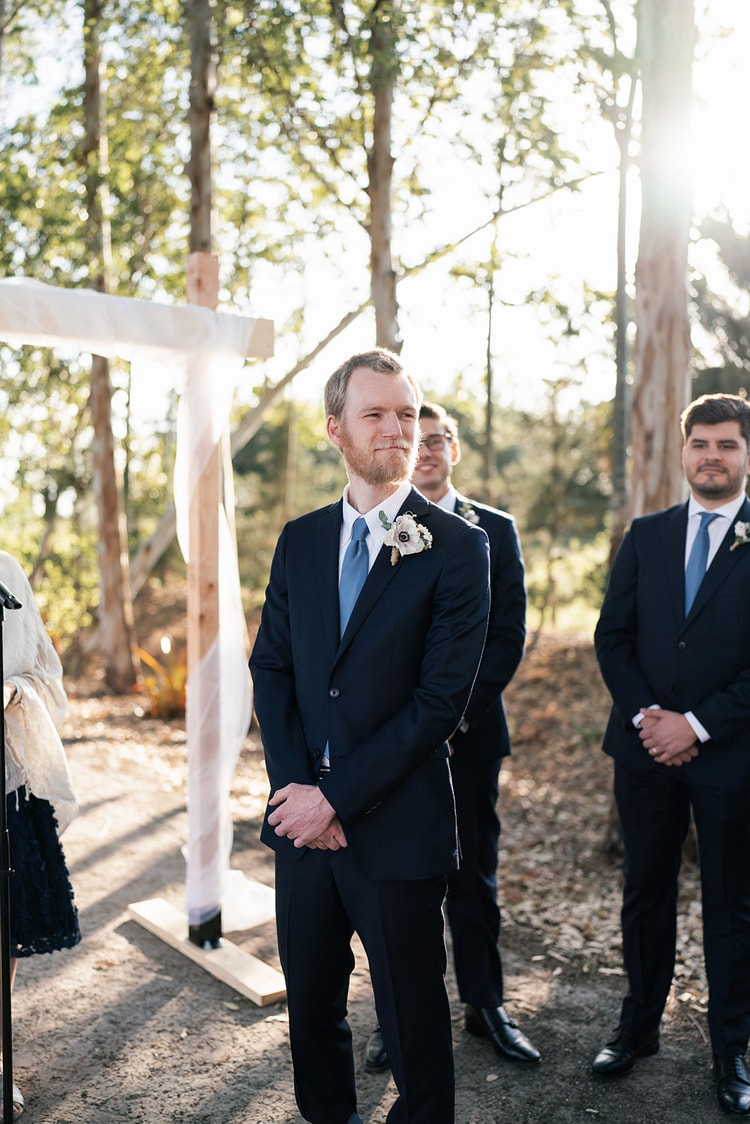 Groom at the altar waiting calmly with a composed smile, groomsmen in navy standing behind him beneath trees glowing in backlit sun.
