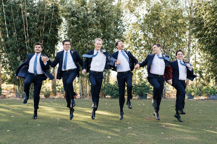 Groom and five groomsmen in navy suits jumping and running across a lawn in front of tall bamboo, full of energy.