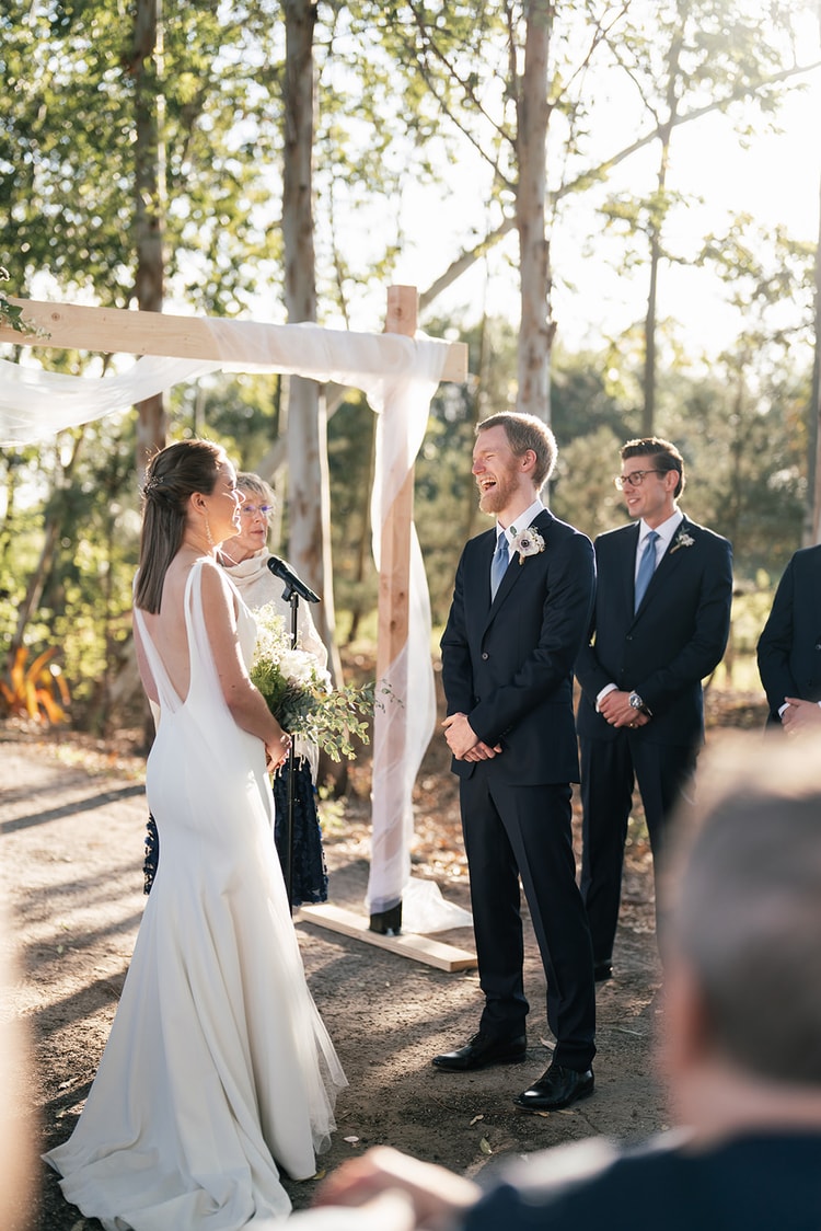 Couple exchanging vows at a wooden ceremony arch draped with white fabric, the officiant beside them and groomsmen watching, in a wooded outdoor setting.