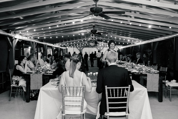 Black-and-white overhead view of the reception room: couple seated at the sweetheart table as a guest delivers a speech to a full room under café lights.