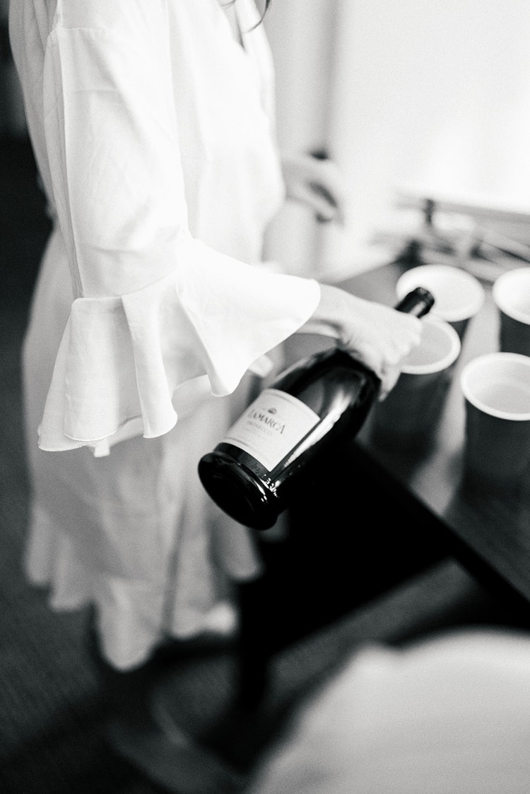 Bride in a ruffled white robe holding a champagne bottle above cups on a table during getting ready, black and white.