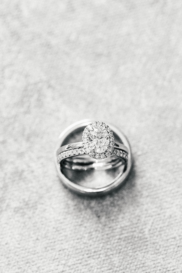 Close-up of a diamond halo engagement ring and two wedding bands stacked together on a soft fabric surface, black and white.