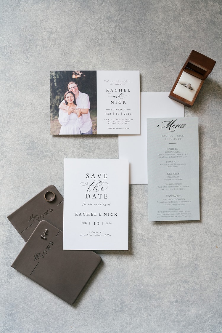 Wedding stationery flat lay featuring save the date, invitation, menu card, vow books, and a diamond ring in a wooden box.