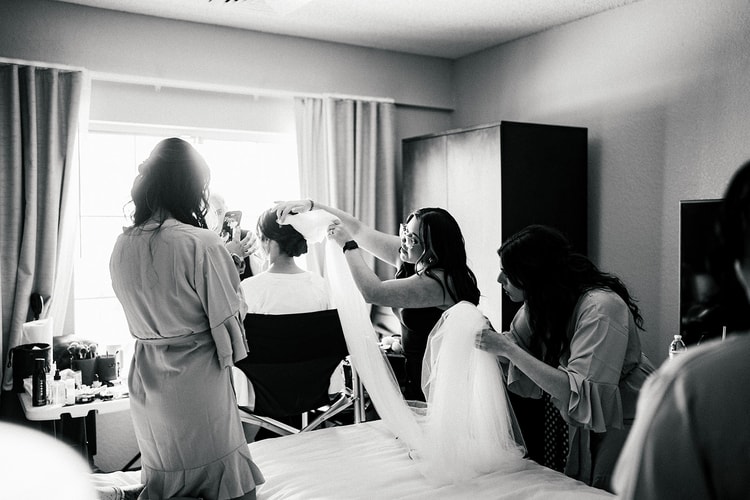 Bridesmaids in silk robes lifting and pinning the bride's cathedral veil during getting ready in a hotel room, black and white.