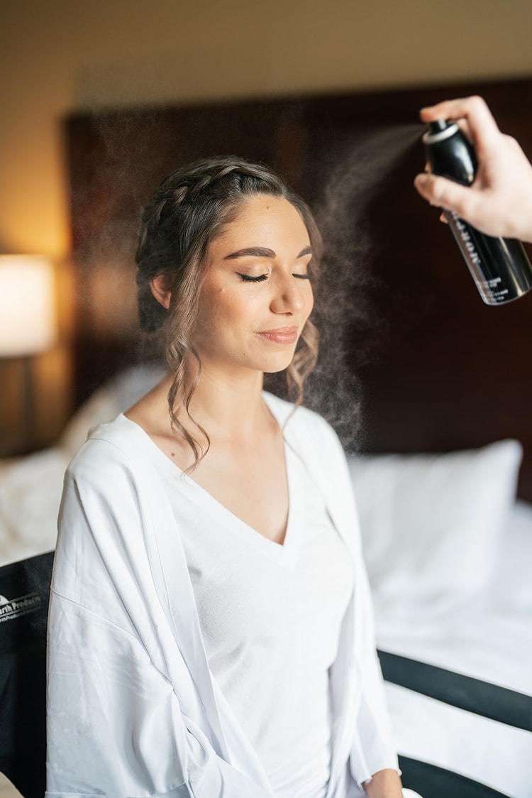 Bride seated in a chair eyes closed as a stylist sprays setting mist over her finished hair and makeup during getting ready.