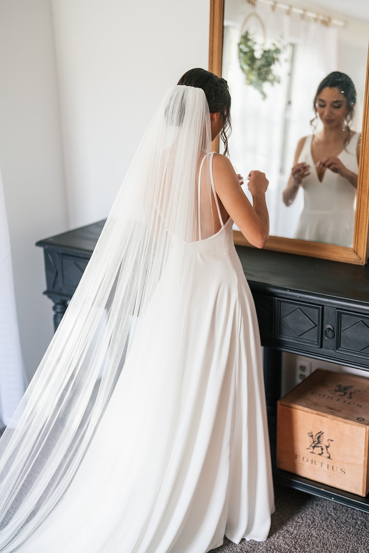 Bride in her gown and cathedral veil looking at her reflection in a gold-framed mirror, adjusting her jewelry before the ceremony.