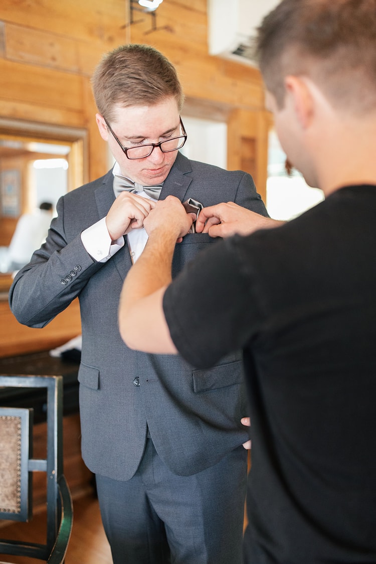 Groomsman pinning a boutonniere on the groom's lapel while the groom adjusts his gray bow tie, getting ready in a wood-paneled room.