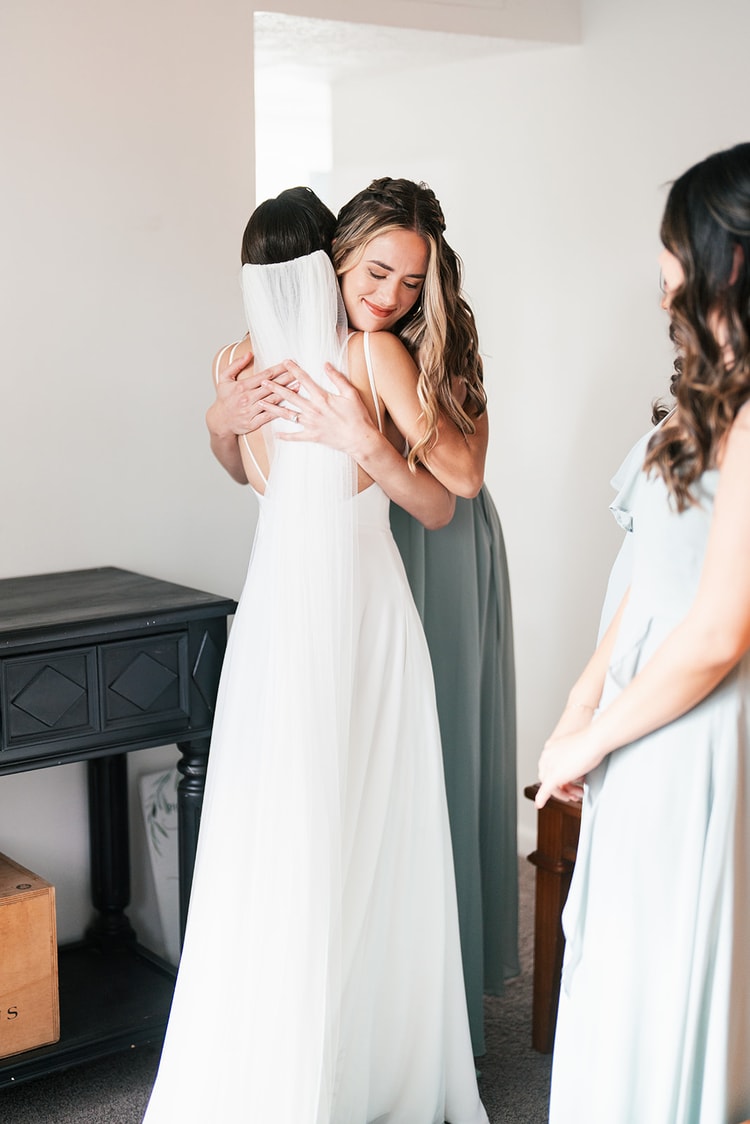 Bridesmaid in a sage green dress hugging the bride tightly during getting ready, both eyes closed, another bridesmaid looking on.