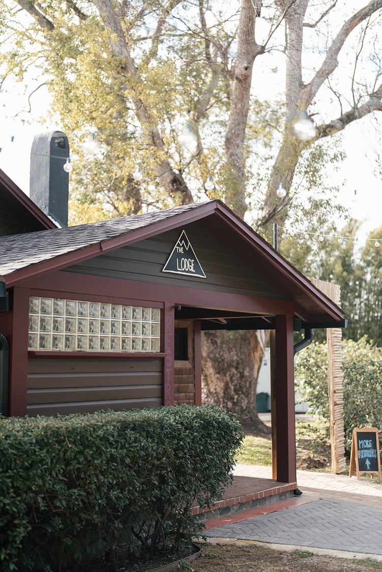 Exterior of a rustic lodge building with a triangular sign at the wedding venue, string lights strung through the trees above.