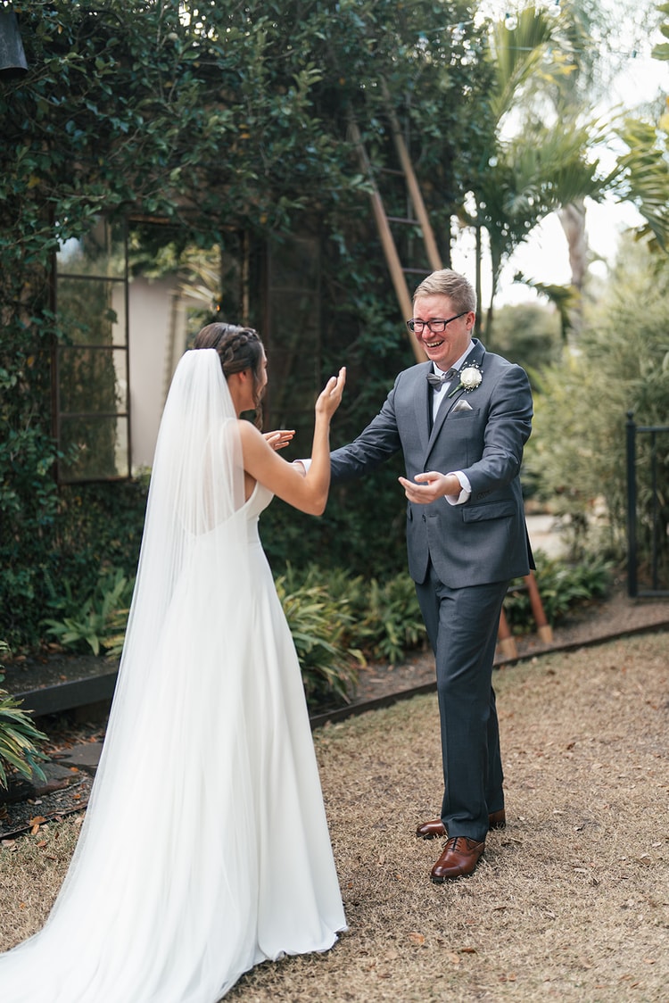 Groom turning toward bride with a wide, joyful laugh during their first look in a lush garden setting.