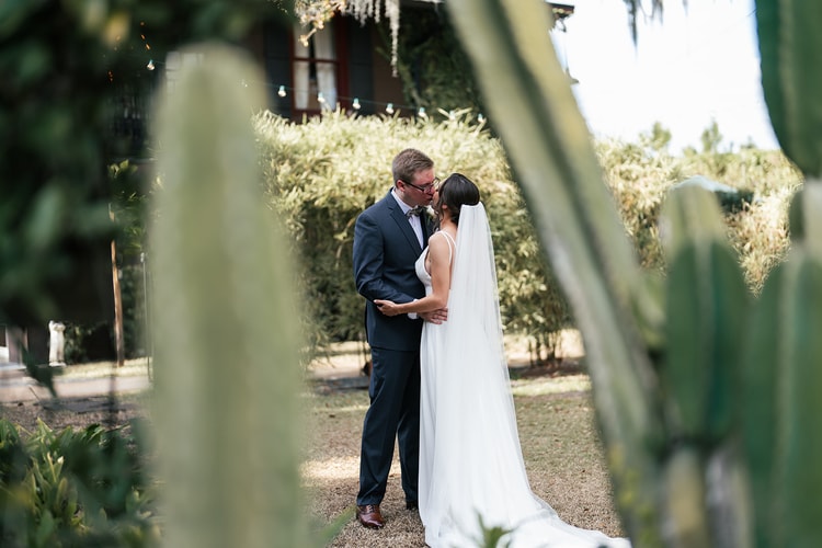 Bride and groom sharing a kiss during their first look, framed between tall cactus and lush garden foliage at the venue.