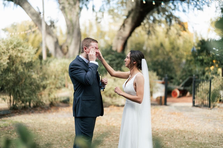 Bride gently wiping a tear from the groom's eye during their emotional first look in the garden, both smiling.