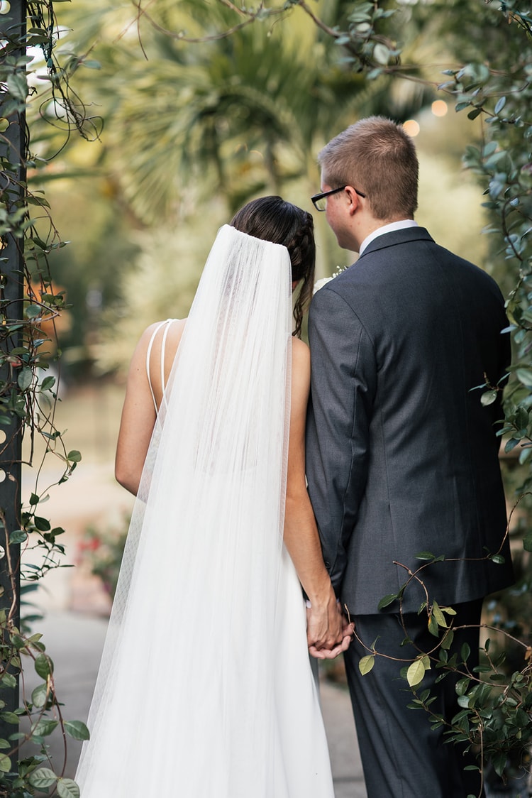 Bride looking over her shoulder at the groom during portraits, a slight smile on her face, lush garden bokeh behind them, black and white.