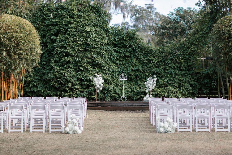 Outdoor ceremony space set with rows of white chairs on either side of a grass aisle, white floral arrangements at the altar end.