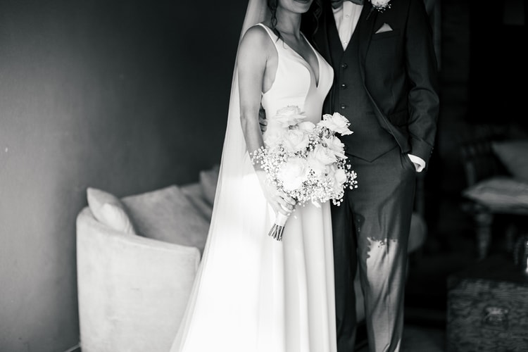 Bride holding her bouquet standing beside the groom in a dimly lit room, a stylized portrait with dramatic contrast, black and white.
